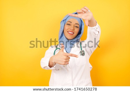 Young beautiful muslim doctor woman wearing medical uniform and hijab making finger frame with hands. Creativity and photography concept. Yellow background