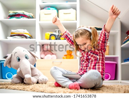 Smiling little girl sitting on the floor and holding her hands up while watching cartoons on tablet