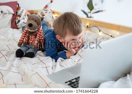 little boy lying in bed in nursery room watching at laptop, distant education, learning new, video chat, watching cartoons, spend time at home. Stay home.