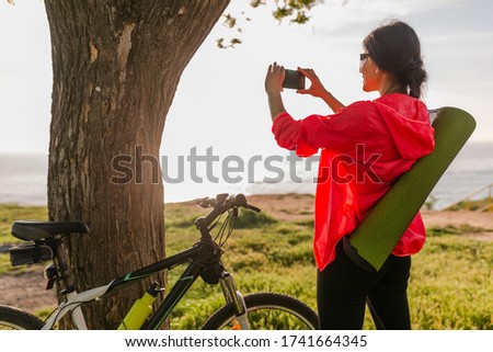slim fit beautiful woman doing sports in morning in park riding on bicycle with yoga mat in colorful fitness outfit, making photo on phone, exploring natures, smiling happy healthy lifestyle