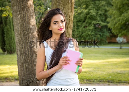 Young beautiful Indian girl stands in the park near a tree, holding a notebook in his hands. Portrait of a female student girl outdoors Royalty-Free Stock Photo #1741720571