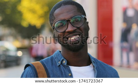 Portrait of attractive african tourist with glasses in blue shirt posing to the camera, smiling with teeth standing in in city center.