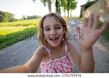 Authentic shot of happy carefree little girl is having fun to make a selfie or video call to parents or friends on a countryside road while taking a walk in a sunny day.