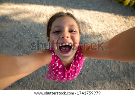 Authentic shot of happy carefree little girl is having fun to make a selfie or video call to parents or friends on a countryside road while taking a walk in a sunny day.