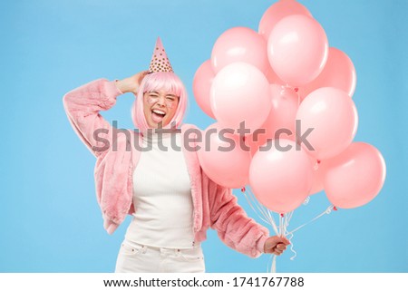 Young birthday woman dressed in pink, holding gelium balloons, winking and touching head with hand as if posing for photo, isolated on blue background