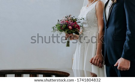 The bride and groom stand at the altar during a wedding ceremony in the church and hold hands. In the bride’s hand is a beautiful bouquet. Empty space for text input.