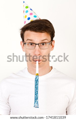 Crop of happy birthday guy posing for picture, with a festive hat and A funny picture of a young guy in white tshirt wearing a festive hat and glasses with the power blowing into a colorful whistle. 