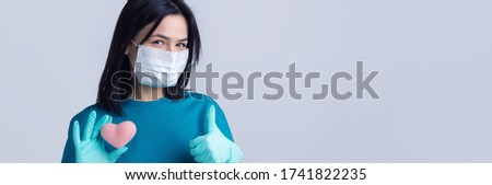 A young brunette girl in a protective medical mask holds a pink heart, symbolizes life.