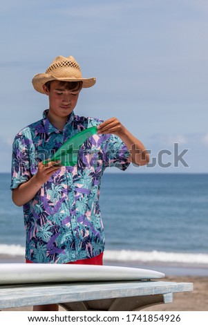 Surfer putting a green fin into a white surfboard on the beach with a blue ocean in the background he is Asian and wearing Hawaiian shirt and straw hat. He is also has a sun tan