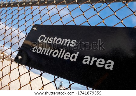 A black metal board sign with the words customs controlled area in white letters. The sign is affixed to a metal mesh wire fence. The blue sky and white fluffy clouds are in the background.