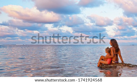 Happy people have fun on summer beach holiday. Young mother with daughter relax at sea water pool. Looking at beautiful view of sunset sky. Healthy family lifestyle, summer travel on tropical island.