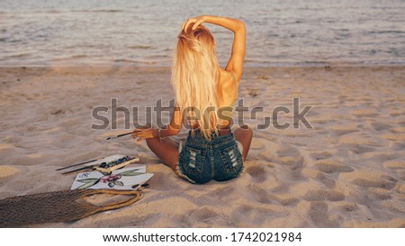 View from back of blond woman with brush looking on the sea, while drawing watercolor picture.