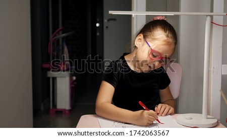 Beautiful girl in glasses draws with felt-tip pen on paper under a lamp in her room