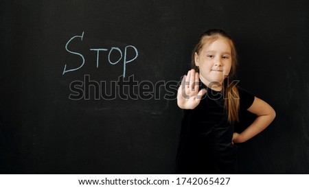 Young beautiful girl of preschool age shows the emotion "Stop". This emotion is also written on a black chalk board behind her back