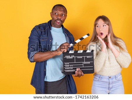 A funny interracial man and woman look scared, horror movies together at home, yellow background
