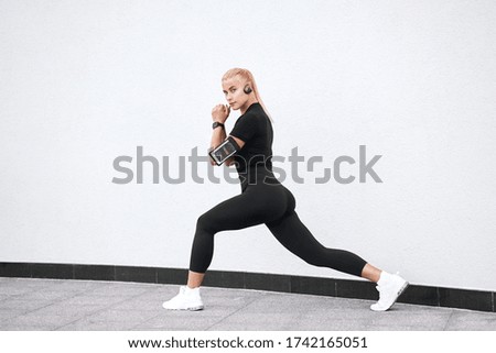 Fitness. Beautiful woman or model wearing black sportswear listening to music in headphones and doing exercises on white urban wall background.                               
