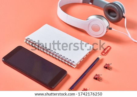 Modern white headphones near smartphone or tablet, blank paper notepad and other stationery things on orange background. Call center concept