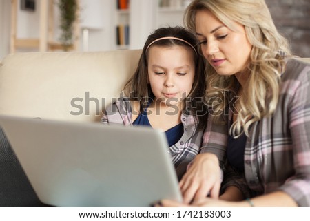 Mother helping little daughter to type on laptop keyboard sitting on the ouch in living room.