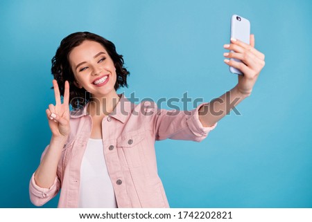 Photo of beautiful cheerful cool lady hold telephone making selfies showing v-sign symbol popular blogger wear casual pink jacket isolated blue color background