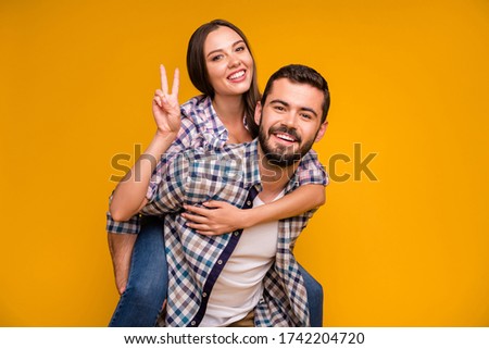 Photo of pretty lady handsome guy couple having fun holding piggyback hands showing v-sign symbol cheerful good mood wear casual plaid shirts jeans isolated yellow color background