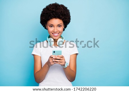 Photo of beautiful dark skin curly lady hipster holding telephone hands choosing song to turn on cool earphones wear casual white t-shirt isolated blue color background