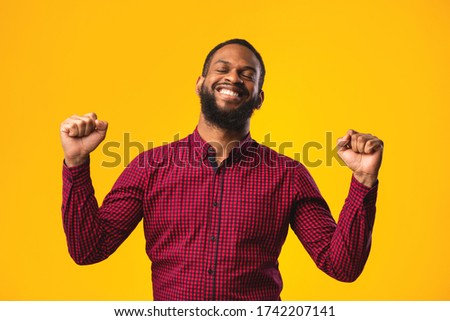 Yes. Excited black guy making winner gesture with clenched fists, smiling with closed eyes over yellow studio wall, copyspace