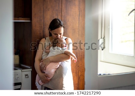Shot of loving mother holding newborn baby,kissing her in head while she is sleeping at home.Young woman standing next to the window,cradling her adorable baby girl at home.Newborn baby in mother arms