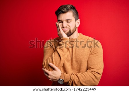 Young blond man with beard and blue eyes wearing casual sweater over red background thinking looking tired and bored with depression problems with crossed arms.