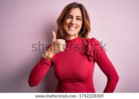 Young beautiful brunette woman wearing casual sweater stnding over pink background doing happy thumbs up gesture with hand. Approving expression looking at the camera showing success.