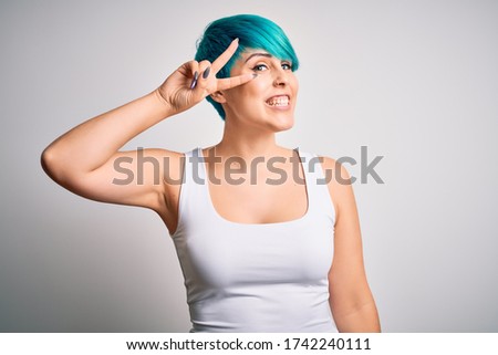 Young beautiful woman with blue fashion hair wearing casual t-shirt over white background Doing peace symbol with fingers over face, smiling cheerful showing victory