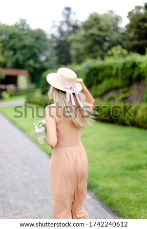 Back view of young blonde woman in body color overalls and hat standing in yeard. Concept of fashion and summer season.