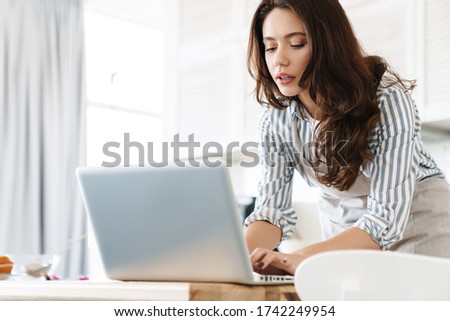 Image of thinking brunette woman wearing apron using laptop while cooking pie in modern kitchen