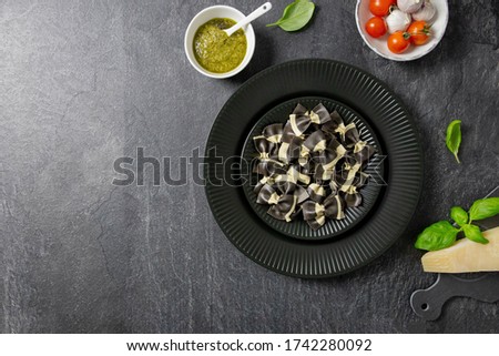 Italian farfalle black pasta, on a dark background stone, black plate. Ingredients for cooking. Cherry Tomatoes, Hard Parmesan, Basil, Pesto, Garlic. Top view. Copy space.