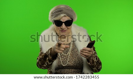 Elderly stylish grandmother caucasian mature woman using smartphone and pointing at camera with hand. Chroma key background. Old granny in fashion wearing browsing on mobile phone. Senior people