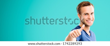 Portrait of happy man in casual smart clothing, showing thumb up gesture, over aqua marine blue color background. Male model at studio picture. Copy space area for some sign text. Wide.