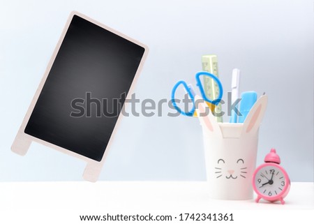 School supplies in paper cup in form of rabbit, pink clock, black chalkboard on white and gray backdrop. Back to school. Stationery. Space for text. 