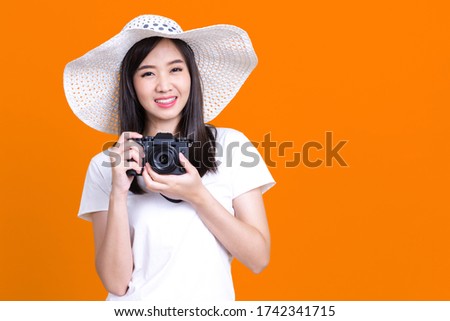 Portrait photo of Asian smiling pretty girl in white shirt wearing beach hat taking photo on camera isolated over orrange background