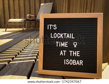Wooden restaurant or pub sign with "locktail time" and "isobar" with a lounge chair in the background, on a wood deck. Felt letter board with white letters. Covid-19 pandemic joke or meme.