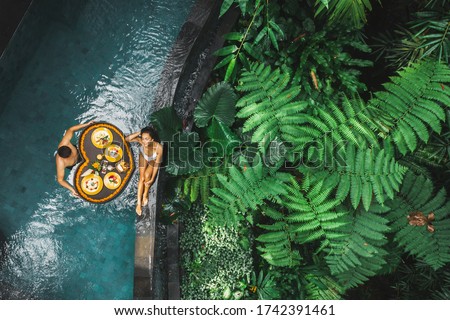 Travel happy couple in love eating floating breakfast in jungle swimming pool. Awakening in morning. Black rattan tray in heart shape, Valentines day or honeymoon surprise, view from above. Royalty-Free Stock Photo #1742391461
