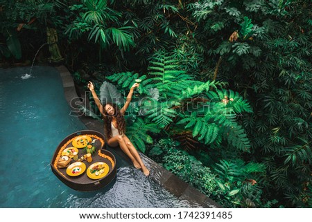 Happy girl relaxing and eating floating breakfast in jungle pool on luxury villa in Bali. Valentines day or honeymoon surprise. Tropical travel lifestyle. Black rattan tray in heart shape.