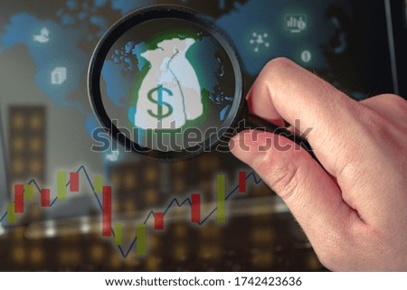 Photo of a hand holding a magnifying glass in front of a laptop screen with a world map with market stock and money symbols.