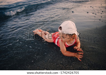 A three-year-old girl in a cap lies on the sand of the sea and looks away. blue color of water