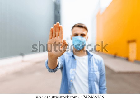 A young man in a protective medical mask makes a stopping hand gesture while standing in the city. Quarantine, epidemic, coronavirus