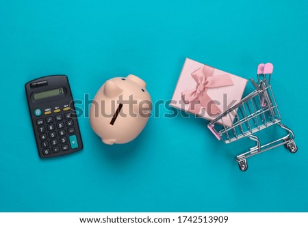 Ready for shopping! Piggy bank and supermarket trolley with gift box, calculator on blue background. Minimalistic studio shot. Overhead view. Flat lay.