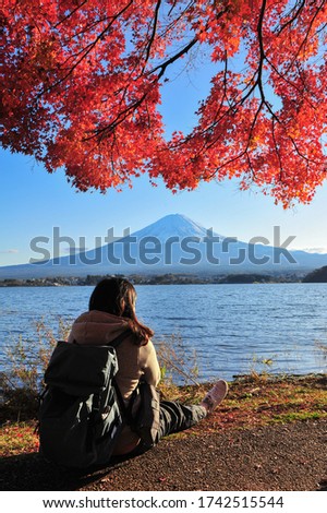 Asian woman backpacker sits and hugs her knees up to the chest under the red maple tree beside lake Kawaguchiko with Fuji mountain background during sunset in autumn