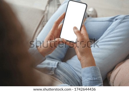 Young woman user customer hold smart phone mock up white screen in hand use mobile shopping app, check social media news, texting mobile sms order food delivery sit on sofa at home. Over shoulder view