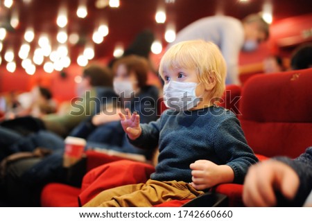 Cute toddler boy wearing face mask watching cartoon movie in the cinema. Lifting virus lockdown. Social distancing restrictions remain. Leisure or entertainment for family with kids after quarantine.