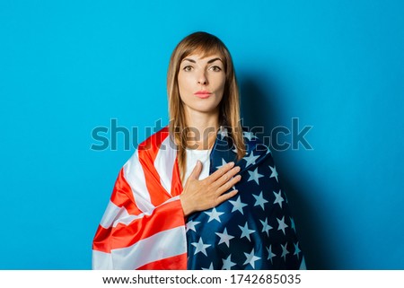 A young woman wrapped in the USA flag makes a hand gesture on a blue background. USA visa concept, English language, gesture is all right, thumbs up, like.