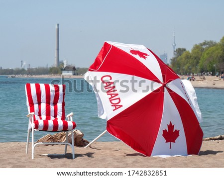 A Canadian flag umbrella and red striped beach chair with the Toronto skyline in the distance celebrating Canada Day on the beach