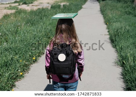 little girl going to school with schoolbag and a book on a head top. girl with backpack outdoors ona pathwalk. back to school concept. learning and education concept. 